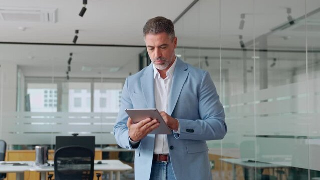 Middle aged business man executive using digital tablet standing in office. Mature older businessman ceo manager wearing suit working on tab analyzing financial trading market data at work.