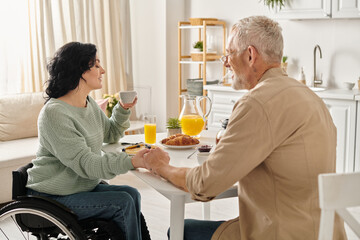A disabled woman in a wheelchair enjoys a moment with her husband at a kitchen table in their home.