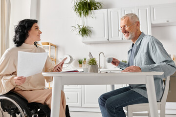 A man in a wheelchair engages in conversation with a woman in a kitchen at home.