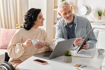 A man and woman, the woman in a wheelchair, sit at a table, engrossed in the screen of a laptop.