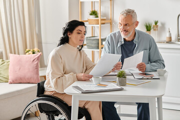 A disabled woman in a wheelchair and her husband sit at a table covered with papers, engaged in a thoughtful discussion.