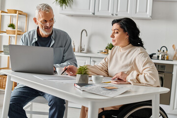 A man and a woman, the woman in a wheelchair, sit together at a table with a laptop, engrossed in...