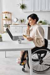 A disabled woman in a wheelchair working remotely from her kitchen, talking on a cell phone.