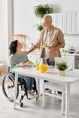 A man in a wheelchair lovingly serves food to his disabled wife in their kitchen at home.