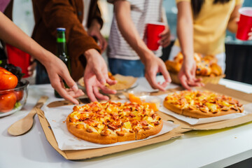 .Young Asian students gather with friends for a pizza party, laughing and sharing slices. Enjoying fast food delivery, they embody diversity and togetherness in a relaxed, enjoyable lifestyle.