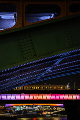 Detail of rivets and the steel structure of a London bridge and a night train running over another bridge illuminated with futuristic purple lights and in the background Tower Bridge and the ship HMS