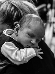 A new born baby boy rests and sleeps on his mothers shoulder. The baby is wearing a three piece suit.