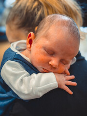 A new born baby boy rests and sleeps on his mothers shoulder. The baby is wearing a three piece suit.
