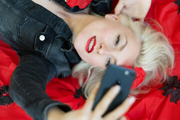 portrait of blonde sensual seductive beauty model with red made up lips and sexy open mouth, with black denim jeans jacket lying in big poppy flowers, takes selfie with cell phone