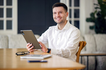 A composed businessman with a genial smile navigates a tablet in a chic, modern workspace.