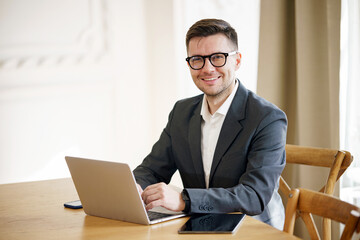 A poised businessman in a suit and glasses works at a laptop, his demeanor reflecting a blend of...