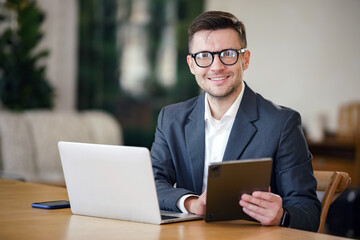 A confident businessman with a pleasant smile multitasks with a tablet and laptop in a...