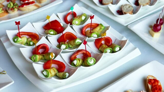 fourchette table with many plates with food.cheese, grape, salami and bread on stick.mini sandwich with salmon, baked pepper and fresh cucumber.many unknown people in background.party, event