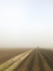 Agricultural field with water furrows being tilled by a tractor driving into the fog