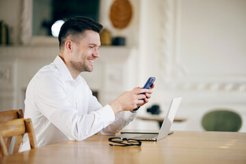 A joyful businessman multitasks with a smartphone and laptop at a stylish wooden desk in a well-lit, elegant space.