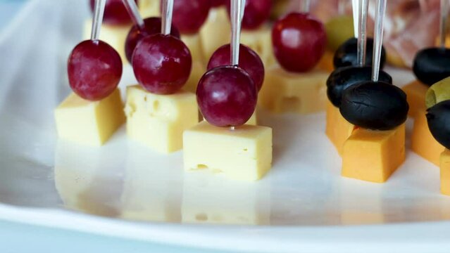 fourchette table with many plates with food.cheese, grape, salami and bread on stick.mini sandwich with salmon, baked pepper and fresh cucumber.many unknown people in background.party, event