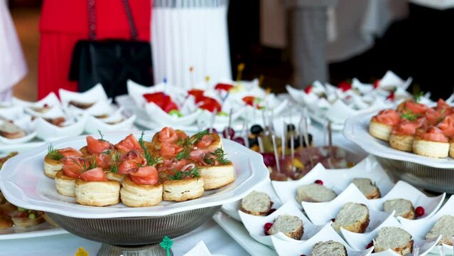 fourchette table with many plates with food.cheese, grape, salami and bread on stick.mini sandwich with salmon, baked pepper and fresh cucumber.many unknown people in background.party, event