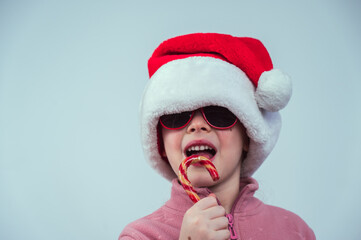 Portrait of a cute Caucasian girl in a Santa Claus hat and sunglasses eating a lollipop on a white background. 