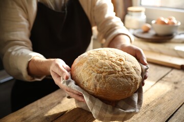Man holding loaf of fresh bread at wooden table indoors, closeup