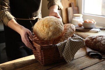 Man holding wicker basket with different types of bread at wooden table indoors, closeup