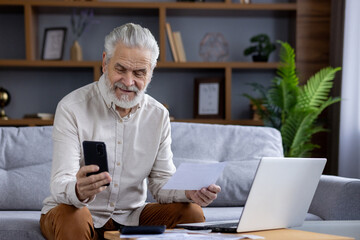 Smiling senior gray-haired man sitting at home on the sofa in front of the table with a laptop,...