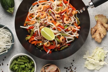 Shrimp stir fry with noodles and vegetables in wok surrounded by ingredients on grey table, flat lay