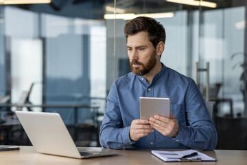 Fototapeta premium A serious young man works in the office at a desk, holds a blanket and looks intently at a laptop screen, compares data and makes an analysis