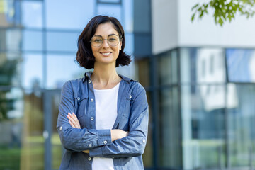 Portrait of a young beautiful woman in casual clothes standing outside an office center and campus, crossing her arms over her chest and looking at the camera with a smile