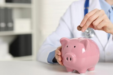 Doctor putting coin into piggy bank at white table indoors, closeup. Space for text