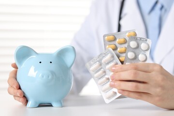Doctor with pills and piggy bank at white table indoors, closeup