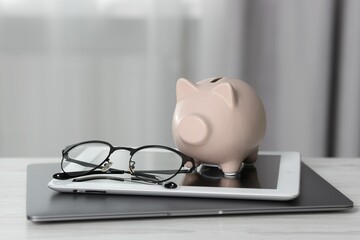 Piggy bank, glasses, tablet and laptop on white table indoors