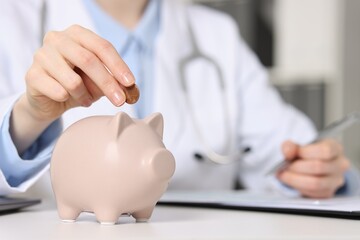 Doctor putting coin into piggy bank while making notes at white table indoors, closeup