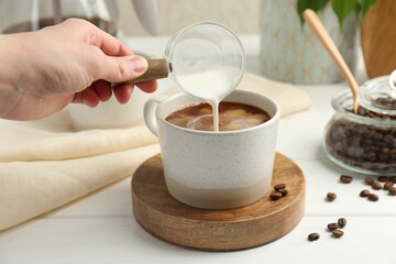 Woman pouring milk into cup with coffee at white wooden table, closeup