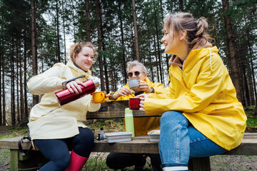 Halt for snack during hiking. Company hikers enjoying picnic on the bench, drinking hot tea, eating sandwiches in forest. Friends relaxing and having snack picnic on nature background. Backpacker trip