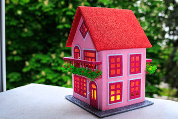 Against a background of green trees, a pink house with a red roof and red light in the windows