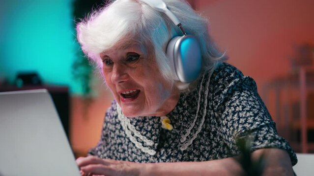 Funny senior woman in headset enjoying gaming competition on laptop, video gamer