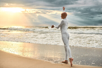 Young hairless ballerina with alopecia in white futuristic suit dancing on seashore at sunset sea,...