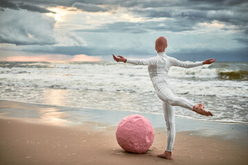 Hairless performer girl with alopecia in white futuristic suit dancing on sea beach with pink...