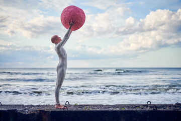 Young hairless girl with alopecia in white futuristic suit standing on concrete fence and holding pink sphere on sea background, metaphoric surreal performance with bald pretty teenage girl