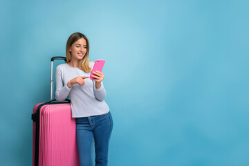  A smiling young lady in casual attire using a mobile phone next to a pink suitcase on a blue background