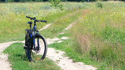 Obraz premium bike stands on the road in the field. A mountain bike stands on a field path with green grass. Mountain bike, blooming summer field, meadow flowers, sunny day. ride a bike. outdoor activities