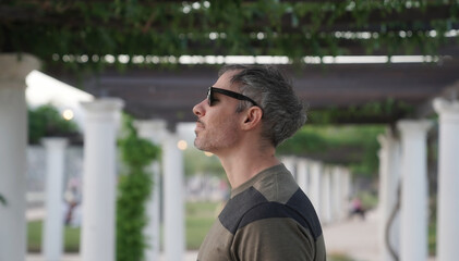 Profile view of a man in his 30s, wearing sunglasses in the park at sunset. The white pergola columns in the background. 