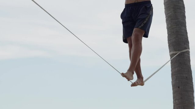 Male walking on the slack rope low angle view. Slack line tightened between palm trees. Healthy lifestyle concept.