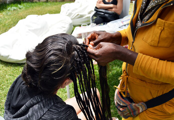 Manos de peluquera haciendo trenzillas africanas en el pelo de una mujer joven al aire libre de...