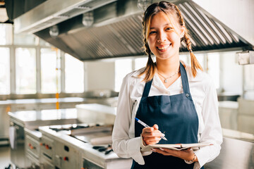 Portrait of a confident woman chef in commercial kitchen. Holds note book teaching with pen....