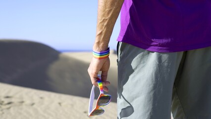 Close up of hand with rainbow bracelet and sunglasses with sand dunes and ocean on the horizon....