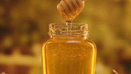 Opened glass jar standing on the table with fresh golden honey, hand dipping honey dipper in the jar with thick syrup, close up shot.