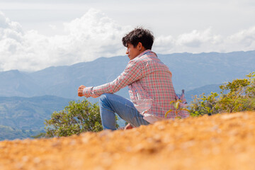 Naklejka premium Lifestyle, young latin man sitting on top of a mountain looking at the panorama in sunny spring and summer morning