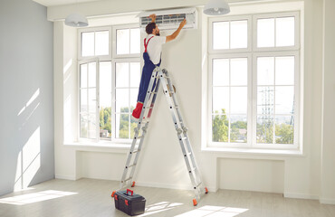 Professional electrician man in uniform standing back on a ladder maintaining or cleaning modern air conditioner indoors. Male technician in the room repairing or installing air conditioner.