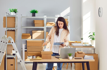 Young confident female employee working in warehouse among cardboard boxes. Woman checking stock and inventory on workplace. Small business owner preparing order for client in storeroom.